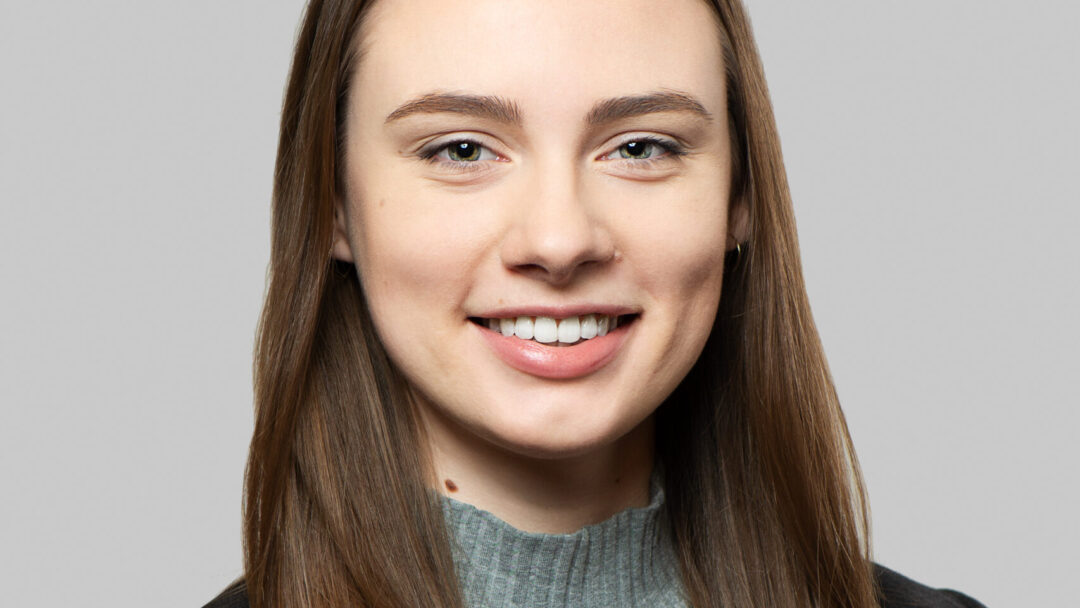 A person wearing a black blazer and a light blue turtleneck sweater against a grey background.