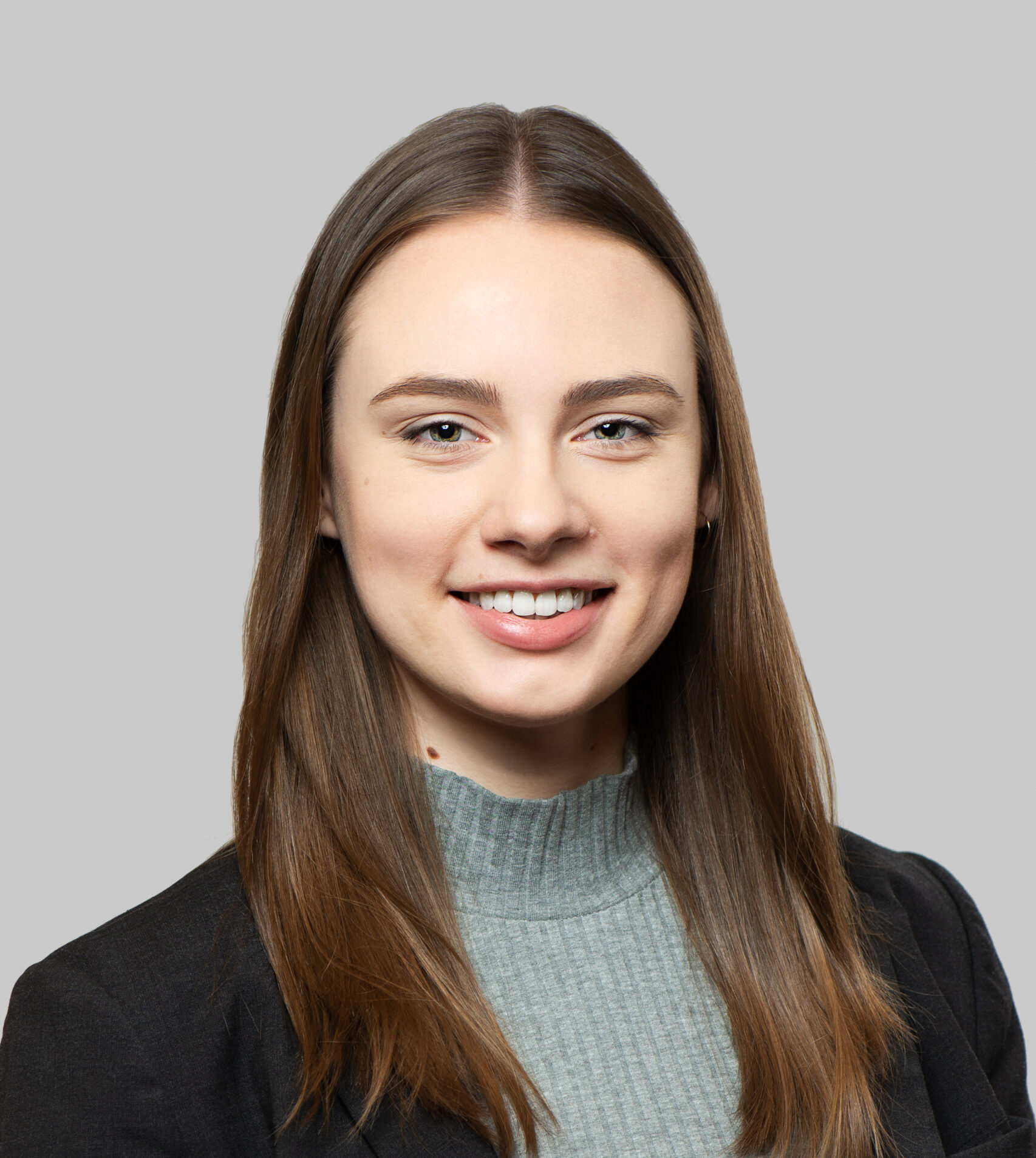 A person wearing a black blazer and a light blue turtleneck sweater against a grey background.