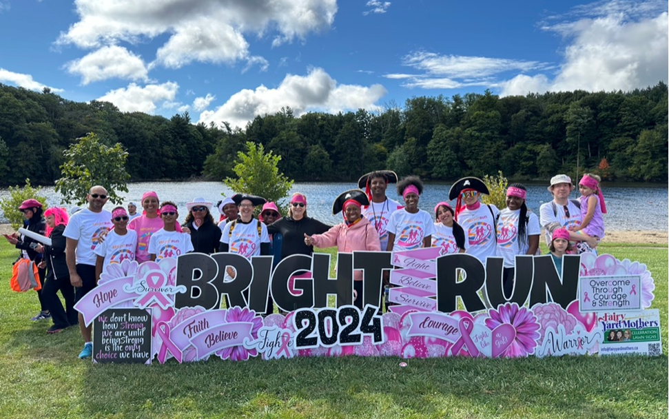 A group of individuals standing behind a large sign for an event called “BRIGHT RUN 2024” in a park setting with trees and a lake in the background. The sign features pink ribbons and various motivational words such as “hope,” “believe,” “courage,” and “fight.” The individuals are wearing event-related attire, with some elements of pink, indicating the event may be related to breast cancer awareness.