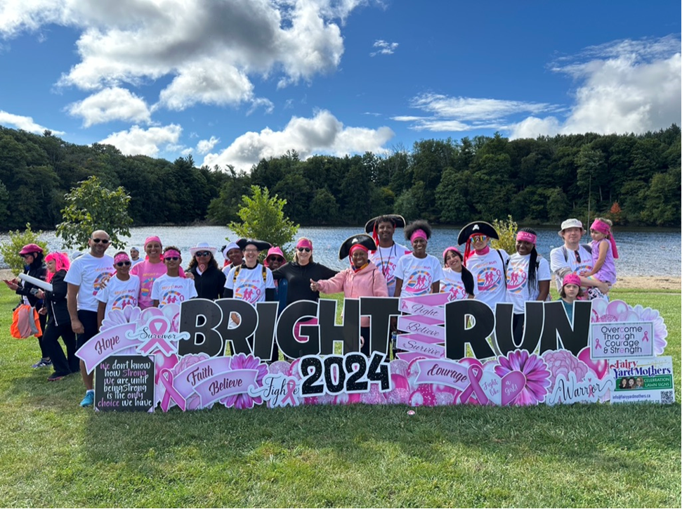 A group of individuals standing behind a large sign for an event called “BRIGHT RUN 2024” in a park setting with trees and a lake in the background. The sign features pink ribbons and various motivational words such as “hope,” “believe,” “courage,” and “fight.” The individuals are wearing event-related attire, with some elements of pink, indicating the event may be related to breast cancer awareness.